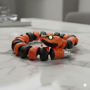 May include: A segmented snake toy in a circular shape. The toy is primarily orange and black, with a silver tail. The snake's head has a green eye and a red tongue. The toy is on a white marble surface.