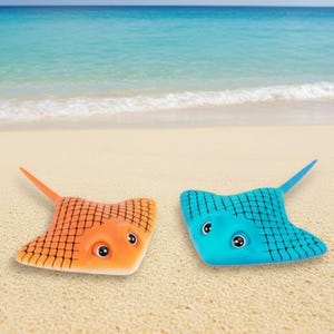 Könnte beinhalten: Zwei Spielzeug-Stachelrochen am Strand. Einer ist orange mit schwarzem Gittermuster, der andere blau. Beide haben große, runde Augen und spitze Schwänze, was ein spielerisches, maritimes Thema erzeugt.
