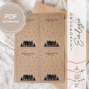 Könnte beinhalten: Ein Bogen mit vier Geschenk-Anhängern aus Kraftpapier mit Weihnachtsmotiv. Jeder Anhänger zeigt eine schwarze Silhouette einer Dorflandschaft mit Bäumen, Sternen und dem deutschen Text "WEIHNACHTEN la ga". Die Worte "PDF DOWNLOAD" stehen in einem Kreis.