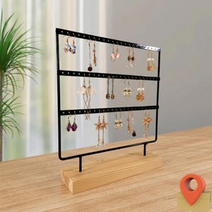 May include: A black metal earring display stand with three horizontal bars, holding various earrings. The stand is mounted on a natural wood base. The earrings are of different styles and colors, including gold, silver, and pearl.
