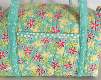 May include: A quilted duffel bag with a floral pattern in red, yellow, and green on a turquoise background. The bag has a front pocket with a white polka dot trim and turquoise handles.