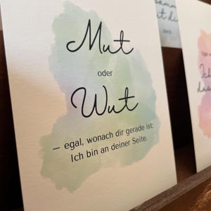 May include: A white card with the German words "Mut oder Wut" in black script, over a watercolor wash of green, blue, and purple. The text "- egal, wonach dir gerade ist: Ich bin an deiner Seite." is below.