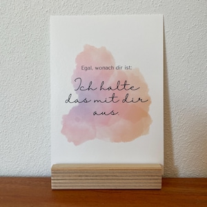 Puede incluir: Una tarjeta blanca con el texto "Egal, wonach dir ist: Ich halte das mit dir aus." se muestra en un soporte de madera. La tarjeta presenta un diseño de estilo acuarela en tonos rosas y morados.