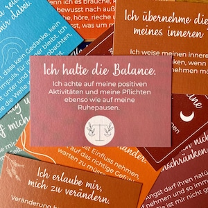 Könnte beinhalten: Eine Sammlung farbenfroher Karten mit inspirierenden Botschaften. Die zentrale Karte, in Rosa, lautet "Ich halte die Balance" (Ich halte die Balance) und zeigt ein Waage-Symbol. Andere Karten haben verschiedene Texte und Farben.