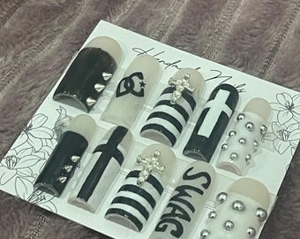 May include: A collection of handmade artificial nails with diverse designs. The nails display black and white patterns, including stripes, crosses, and the word "SWAG." Some nails are decorated with silver studs and a cross-shaped jewel.