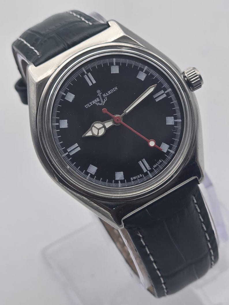 K&ouml;nnte beinhalten: Eine Ulysse Nardin Armbanduhr aus Edelstahl mit schwarzem Zifferblatt, wei&szlig;en Stundenmarkierungen und einem roten Sekundenzeiger. Die Uhr hat ein schwarzes Lederarmband mit wei&szlig;en N&auml;hten. Das Zifferblatt zeigt den Markennamen und "SWISS MADE".