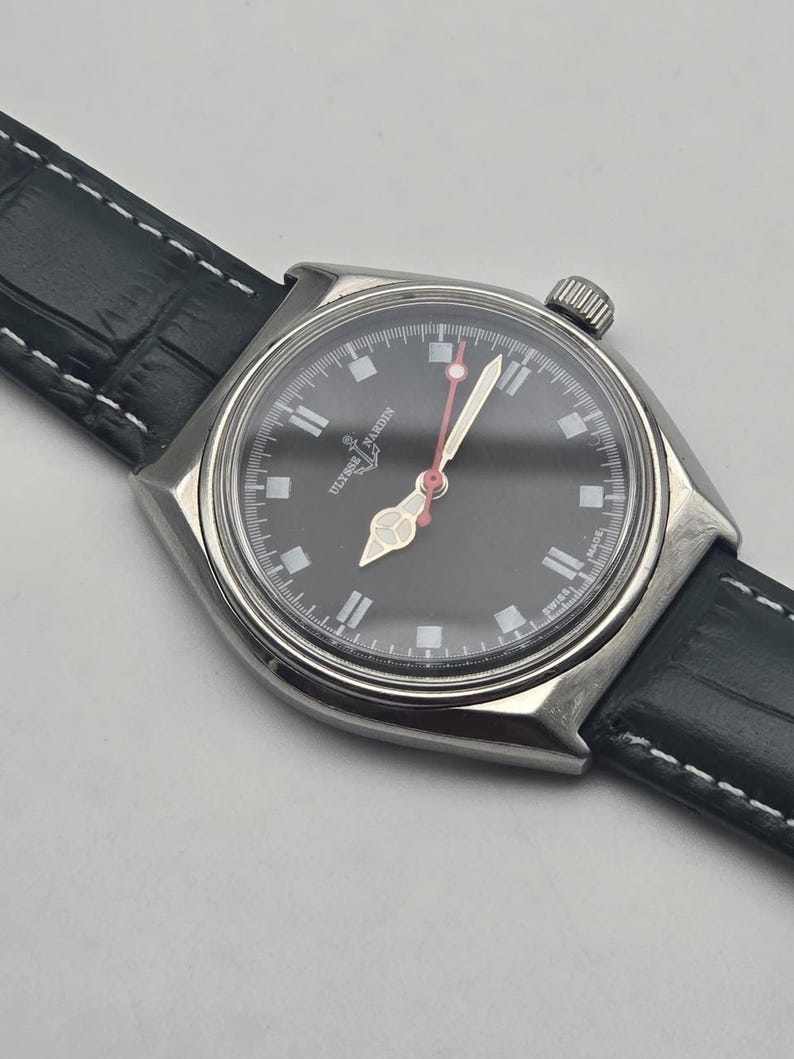 K&ouml;nnte beinhalten: Eine Edelstahl-Armbanduhr mit schwarzem Zifferblatt und schwarzem Lederarmband. Das Zifferblatt der Uhr hat wei&szlig;e Stundenmarkierungen und Zeiger, mit einem roten Sekundenzeiger. Die Uhr ist mit "SWISS MADE" gekennzeichnet.