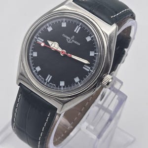 K&ouml;nnte beinhalten: Eine Ulysse Nardin Uhr mit schwarzem Zifferblatt und schwarzem Lederarmband. Die Uhr hat ein silbernes Geh&auml;use, wei&szlig;e Stundenmarkierungen und rote Akzente auf den Zeigern. Das Armband ist aus schwarzem Leder mit wei&szlig;en N&auml;hten.