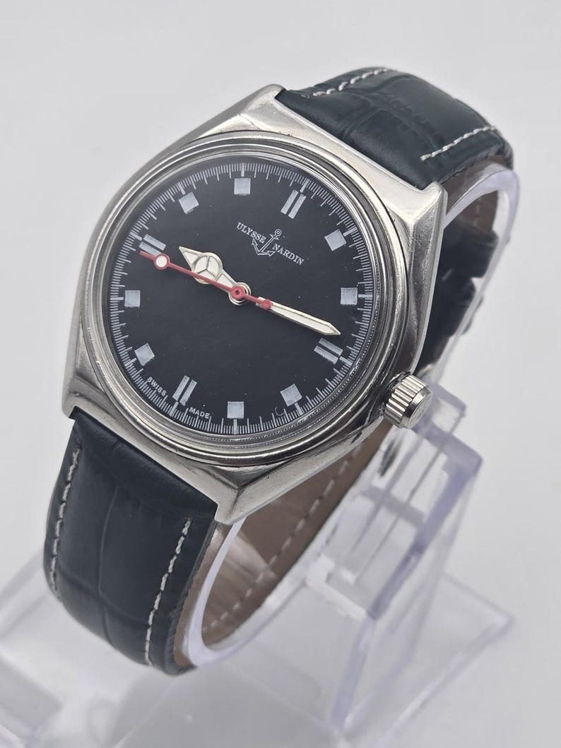 K&ouml;nnte beinhalten: Eine Ulysse Nardin Uhr mit schwarzem Zifferblatt und schwarzem Lederarmband. Die Uhr hat ein silbernes Geh&auml;use, wei&szlig;e Stundenmarkierungen und rote Akzente auf den Zeigern. Das Armband ist aus schwarzem Leder mit wei&szlig;en N&auml;hten.