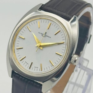 May include: A vintage Ulysse Nardin wristwatch with a silver-colored case and a white face. The watch features gold-colored hands and markers, and a black leather band. The watch is marked "Swiss Made".