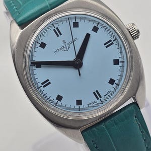 Pu&ograve; includere: Un orologio Ulysse Nardin vintage con quadrante azzurro e lancette e indici neri. L'orologio ha una cassa argentata e un cinturino in pelle verde acqua. Il quadrante mostra il logo Ulysse Nardin e la scritta "SWISS MADE".