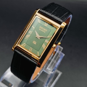 Puede incluir: Un reloj Seiko Quartz rectangular dorado con esfera verde y correa de cuero negro. La esfera del reloj presenta números romanos y agujas doradas. El reloj se exhibe sobre un soporte transparente sobre un fondo oscuro.