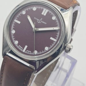 May include: A silver-toned wristwatch with a burgundy face and brown leather band. The watch face features white hour markers and hands, with the brand name "Ulysse Nardin" visible. The watch is displayed on a clear stand.