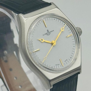 May include: A silver-toned Ulysse Nardin wristwatch with a light gray face and gold hands and markers. The watch features a black leather band and the brand name and logo are visible on the face. The watch is marked "Swiss Made".