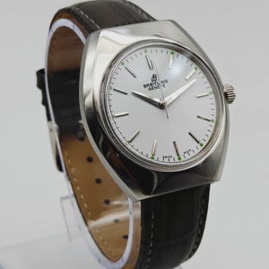 May include: A vintage Breitling Geneve watch with a silver-toned case and a white dial. The watch features a black leather band and a silver crown. The dial has silver hands and green markers. The watch is a classic timepiece.