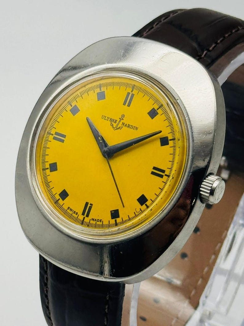 May include: A vintage Ulysse Nardin wristwatch with a bright yellow face and black hands. The watch has a silver-colored case and a dark brown leather strap. The watch face features black rectangular hour markers and the brand name.