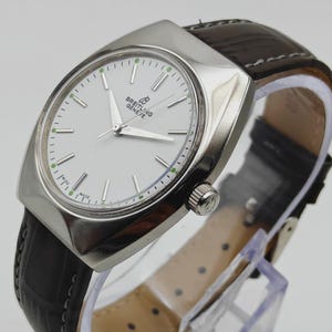 May include: A silver Breitling Geneve watch with a white face and a dark brown leather band. The watch has a rectangular case and a classic design. The watch band is approximately 8 inches long.