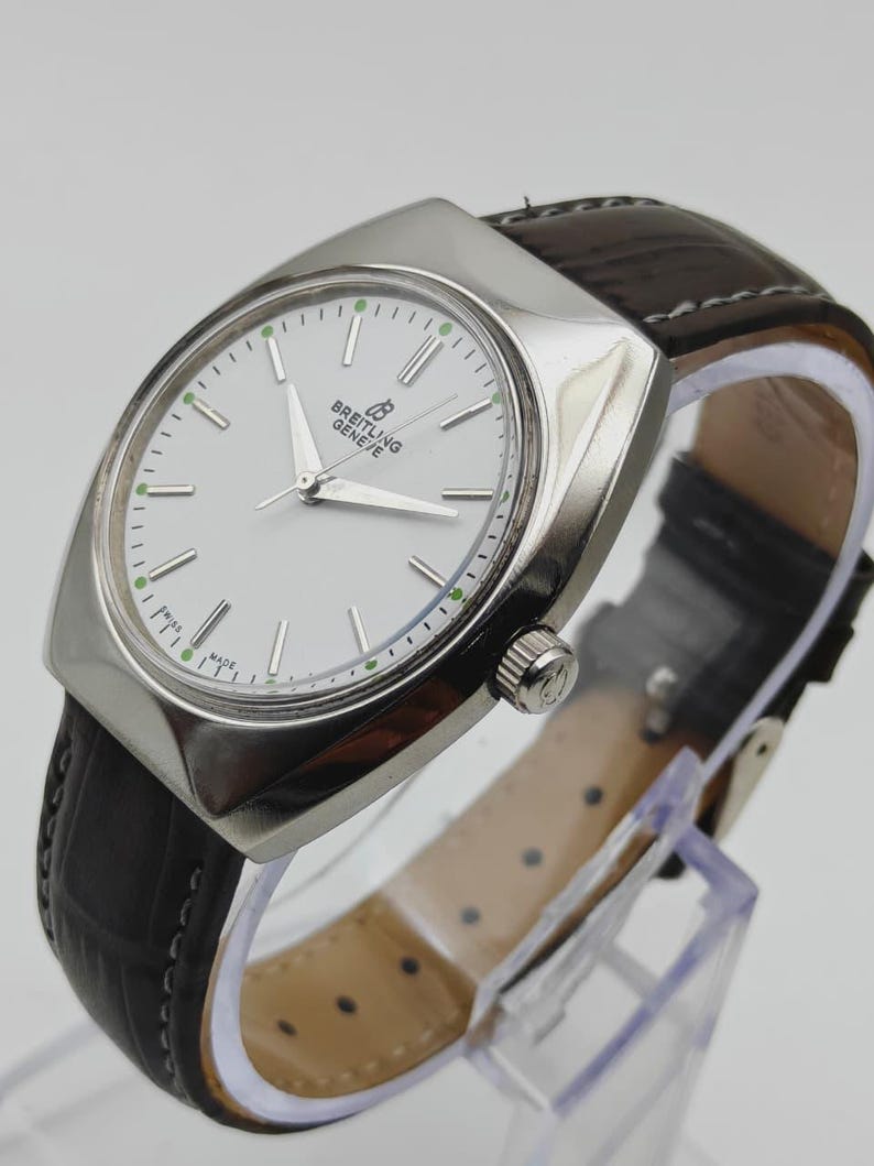 May include: A silver Breitling Geneve watch with a white face and a dark brown leather band. The watch has a rectangular case and a classic design. The watch band is approximately 8 inches long.