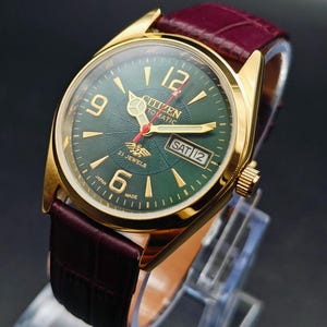 May include: A gold-plated Citizen automatic watch with a green dial and a burgundy leather strap. The watch face shows the day and date, with gold hour markers and hands. The watch is marked "Citizen Automatic" and "21 Jewels".