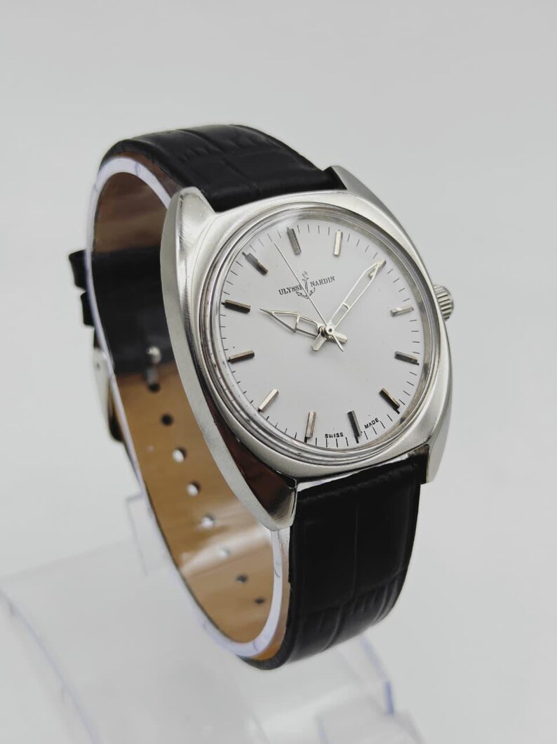 May include: A silver Ulysse Nardin wristwatch with a white face and black leather strap. The watch has a round face with silver hands and markers. The strap is textured and the watch is displayed on a clear stand.