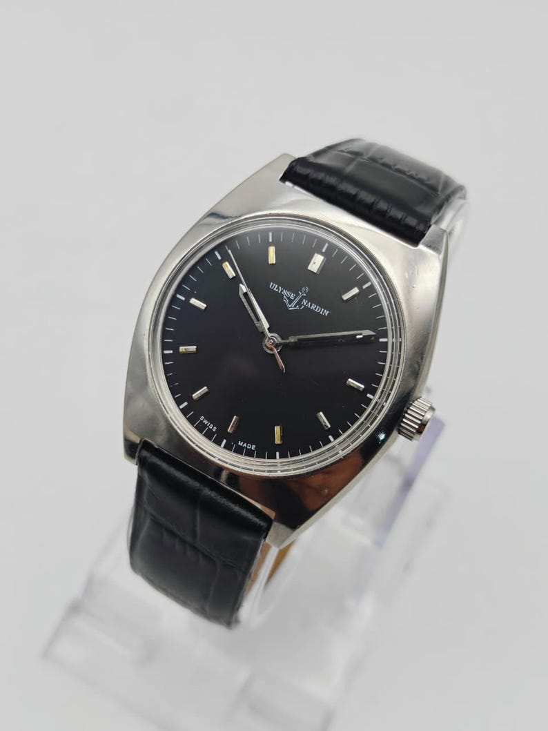 May include: A silver-toned Ulysse Nardin wristwatch with a black face and black leather band. The watch has a rectangular case and a classic design. The watch face features silver hour markers and hands. The band is made of black leather with a crocodile pattern.