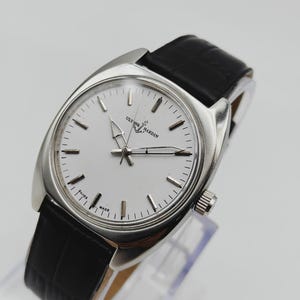 May include: A silver Ulysse Nardin wristwatch with a white face and black leather strap. The watch has a round face with silver hour markers and hands. The strap is made of black leather.