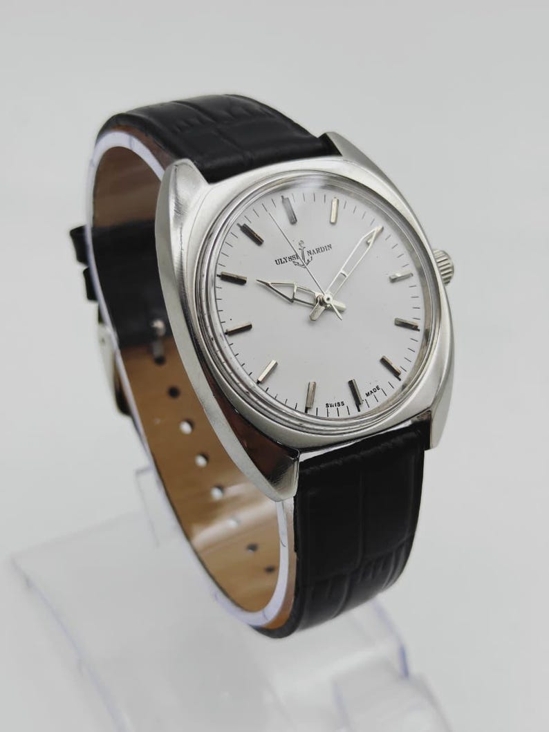 May include: A silver wristwatch with a white face and a black leather strap. The watch face displays the text "ULYSSES NARDIN SWISS MADE". The strap has a crocodile skin pattern.