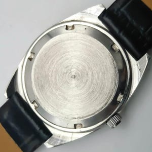 May include: A silver-toned wristwatch with a black leather band. The watch case back is visible, showing a circular brushed metal design. The watch has a small crown and the band is attached with spring bars.