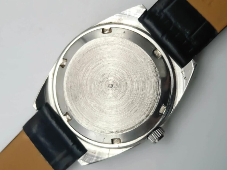 May include: A silver-toned wristwatch with a black leather band. The watch case back is visible, showing a circular brushed metal design. The watch has a small crown and the band is attached with spring bars.