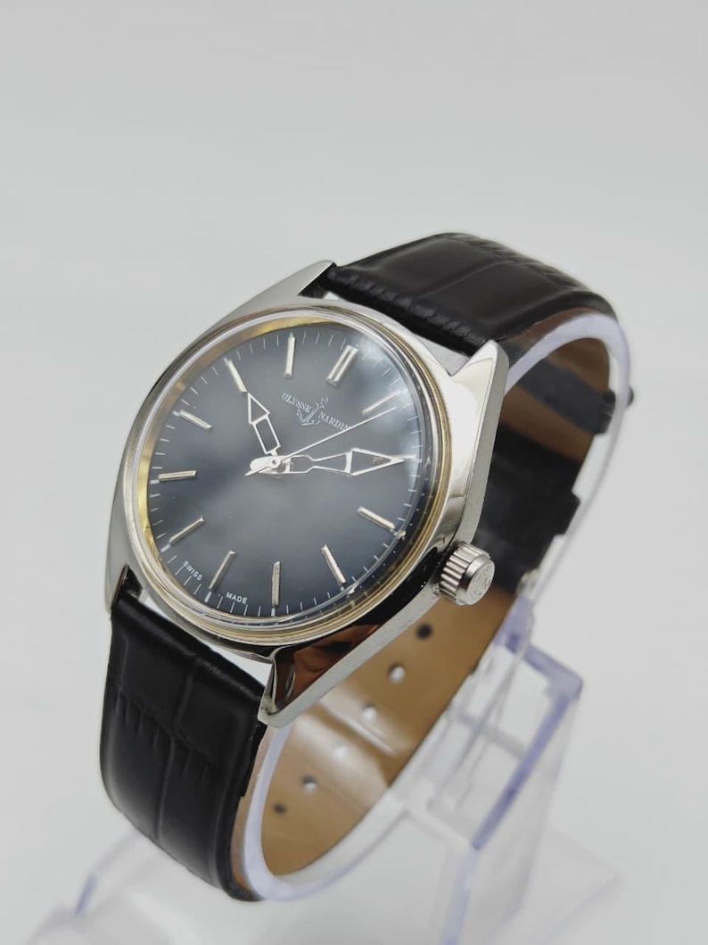 Puede incluir: Un reloj de pulsera plateado con esfera gris oscuro y correa de cuero negro. El reloj presenta marcadores de hora y agujas plateadas, y el nombre de la marca es visible en la esfera. La correa tiene un patr&oacute;n texturizado similar al de un cocodrilo.
