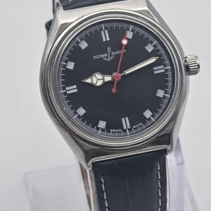K&ouml;nnte beinhalten: Eine Ulysse Nardin-Uhr aus Edelstahl mit schwarzem Zifferblatt, wei&szlig;en Markierungen und einem roten Sekundenzeiger. Die Uhr hat ein Lederarmband mit wei&szlig;en N&auml;hten. Die Uhr wird auf einem transparenten St&auml;nder pr&auml;sentiert.
