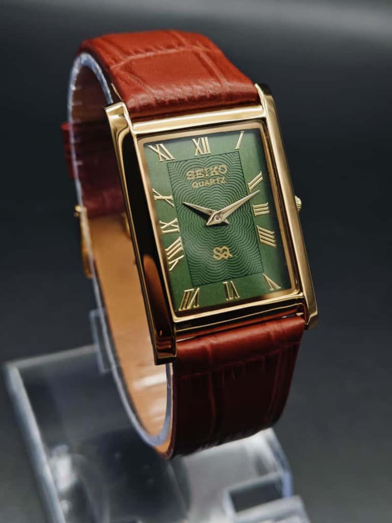 Puede incluir: Un reloj Seiko Quartz dorado rectangular con esfera verde y n&uacute;meros romanos. El reloj tiene agujas doradas y una correa de cuero marr&oacute;n. El reloj se exhibe sobre un soporte transparente.