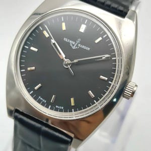 May include: A silver-toned Ulysse Nardin watch with a black face and a black leather band. The watch face has silver and gold-toned markers and hands. The watch is marked "SWISS MADE".