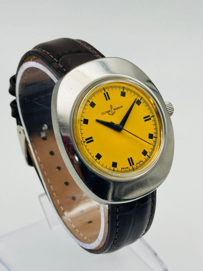 May include: A vintage Ulysse Nardin wristwatch with a bright yellow face and black markers. The watch has a silver-tone case and a dark brown leather band. The watch face displays the brand name and the words "SWISS MADE".