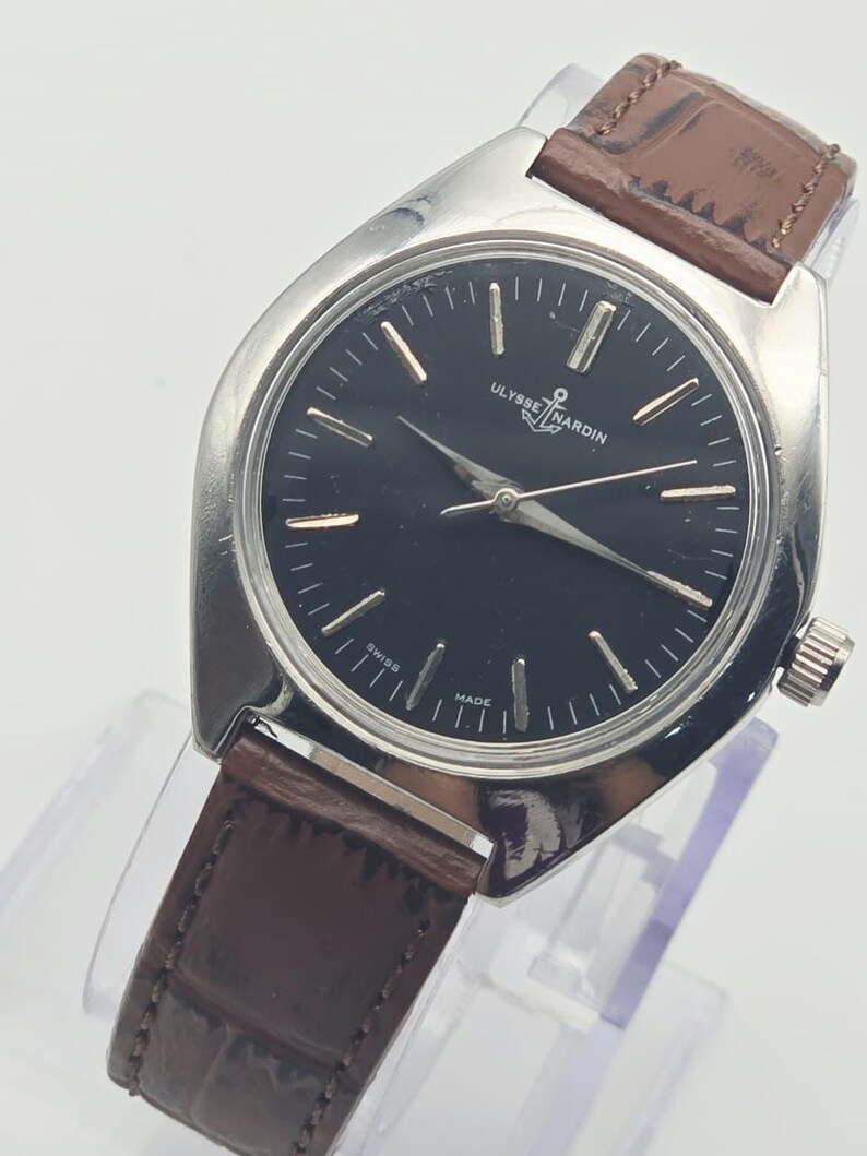 May include: A silver-toned Ulysse Nardin watch with a black face and brown leather band. The watch features gold-colored hour markers and hands. The band has a crocodile pattern. The watch is displayed on a clear stand.