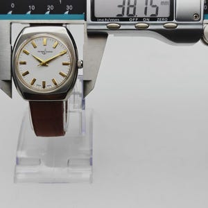 K&ouml;nnte beinhalten: Eine silberfarbene Uhr mit wei&szlig;em Zifferblatt, goldenen Zeigern und Markierungen sowie einem braunen Lederarmband. Die digitale Messschieberanzeige zeigt einen Durchmesser von 38,15 mm. Die Uhr befindet sich auf einem transparenten Kunststoffst&auml;nder.