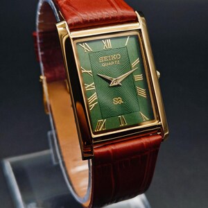 Puede incluir: Un reloj Seiko Quartz rectangular dorado con esfera verde y n&uacute;meros romanos dorados. El reloj tiene una correa de cuero marr&oacute;n. La esfera del reloj tiene el logotipo de Seiko Quartz. El reloj se exhibe sobre un soporte transparente.