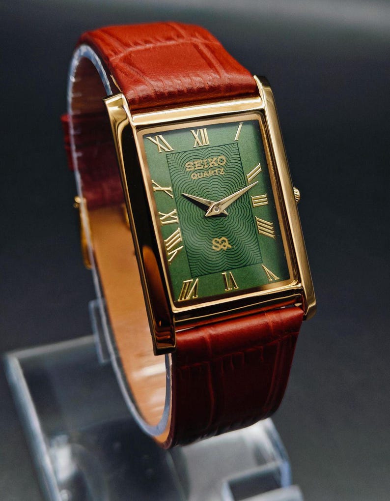 Puede incluir: Un reloj Seiko Quartz rectangular dorado con esfera verde y n&uacute;meros romanos dorados. El reloj tiene una correa de cuero marr&oacute;n. La esfera del reloj tiene el logotipo de Seiko Quartz. El reloj se exhibe sobre un soporte transparente.