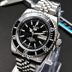 May include: A silver-tone Seiko 5 Sports automatic wristwatch with a black dial, a rotating black bezel with white numerals, and a stainless steel link bracelet. The dial displays the Seiko logo, day-date window, and text reading WATER 10 BAR RESIST.