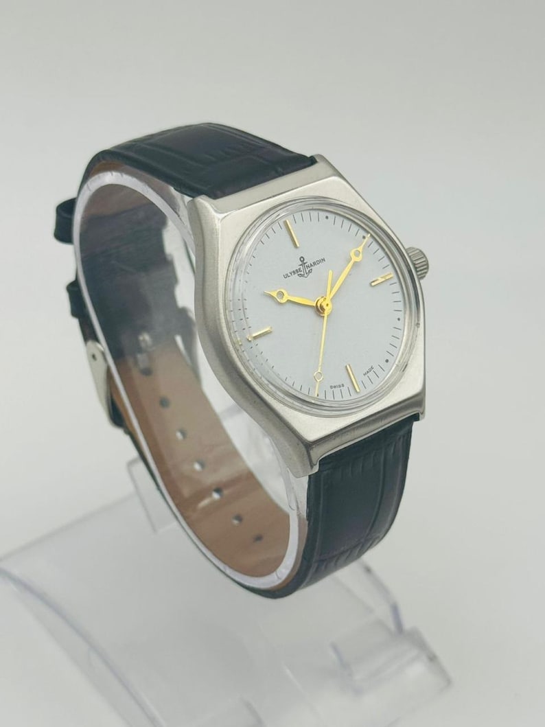 May include: A silver-toned wristwatch with a white face and gold hands and markers. The watch has a black leather band. The watch face has the words "ULYSSSE NARDIN" printed on it.