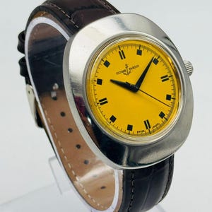 May include: A vintage Ulysse Nardin wristwatch with a bright yellow face and black markers. The watch has a silver-tone case and a dark brown leather band. The watch face displays the brand name and a small anchor logo.