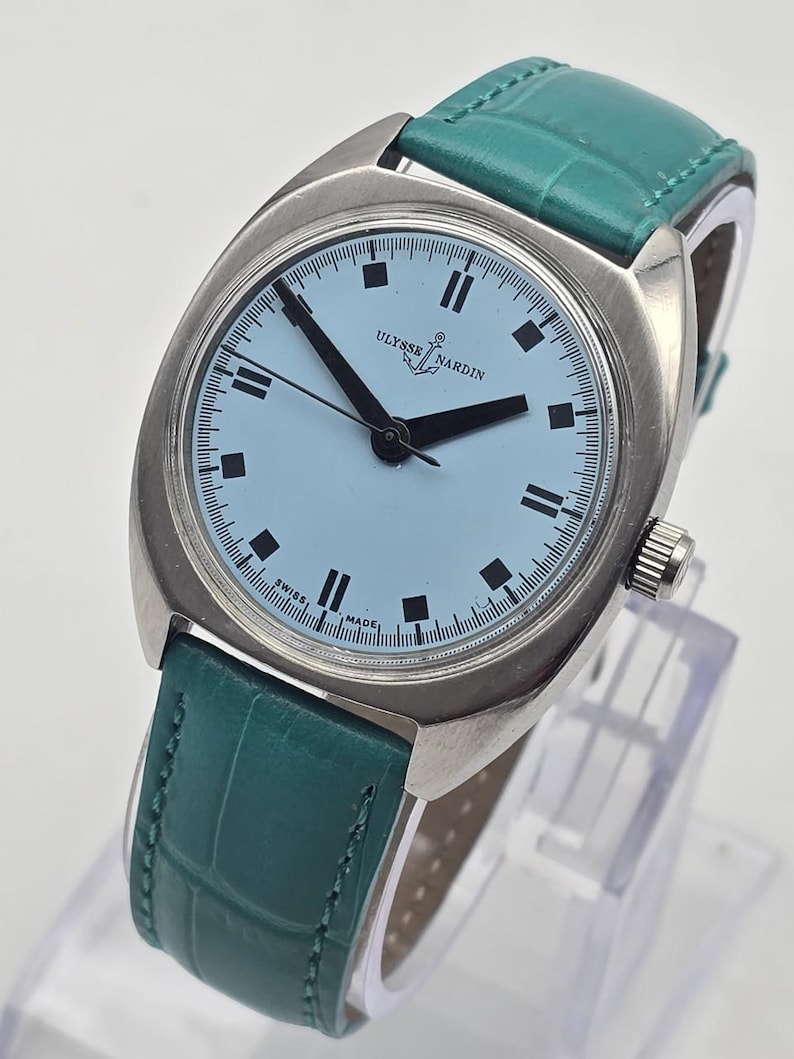 Pu&ograve; includere: Un orologio da polso argentato con quadrante azzurro e lancette nere. L'orologio presenta la scritta "ULYSSE NARDIN" e un cinturino in pelle verde. Il quadrante ha indici delle ore rettangolari neri.