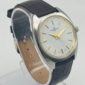 May include: A silver-toned wristwatch with a white face and gold hands. The watch features the brand name "ULYSSE NARDIN" and a small anchor logo. It has a dark brown leather band.