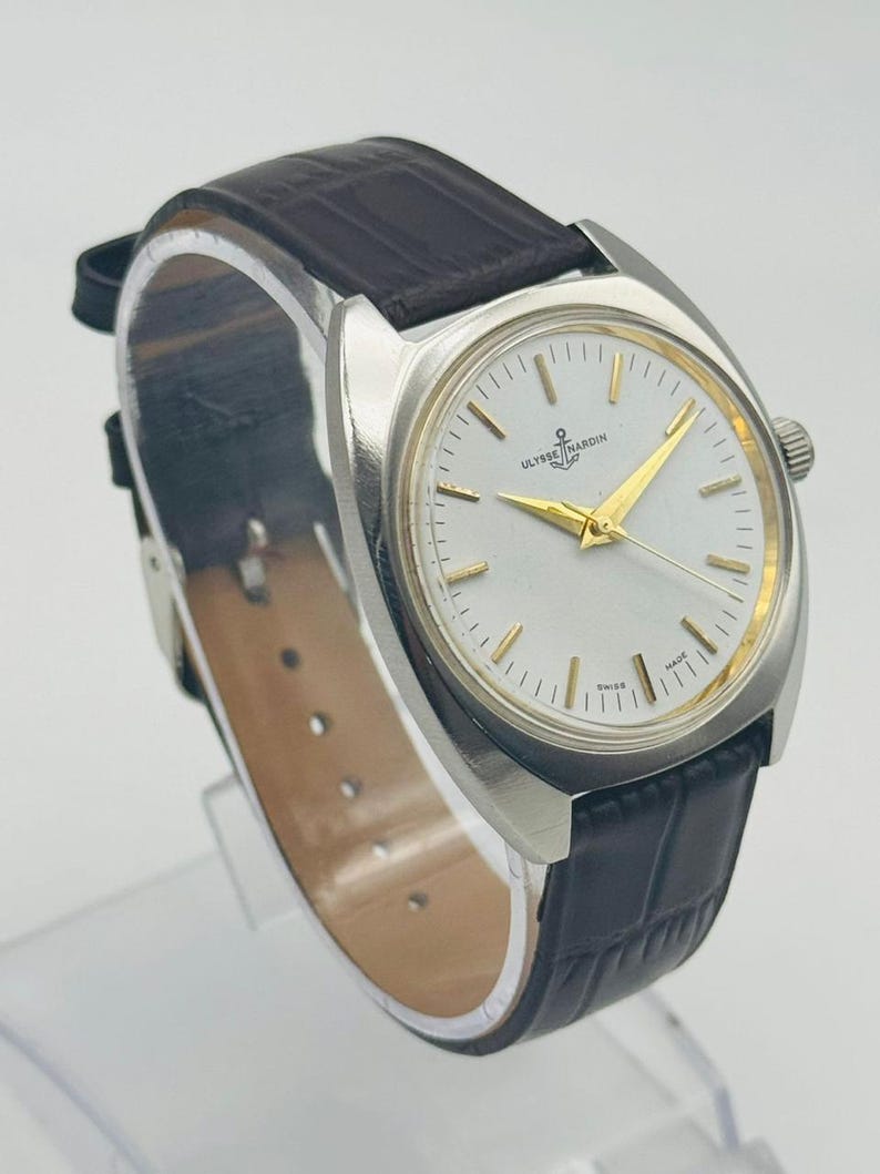 May include: A silver-toned wristwatch with a white face and gold hands. The watch features the brand name "ULYSSE NARDIN" and a small anchor logo. It has a dark brown leather band.