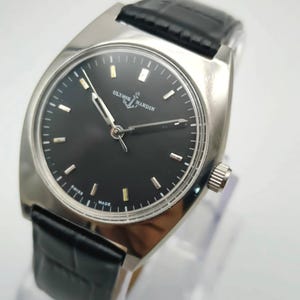 May include: A silver-toned wristwatch with a black face and a black leather band. The watch face has silver-toned hour markers and hands. The watch is marked "Ulysse Nardin" and "Swiss Made".