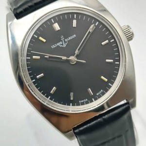 May include: A silver-toned Ulysse Nardin wristwatch with a black face and a black leather band. The watch has a round face with silver and gold-toned markers and hands. The watch face has the words "Ulysse Nardin" and "Swiss Made" printed on it.