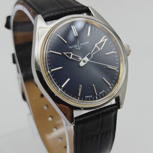 Puede incluir: Un reloj de pulsera Ulysse Nardin plateado con esfera azul oscuro y correa de cuero negro. El reloj presenta agujas e indicadores plateados, con las palabras "SWISS MADE" visibles en la esfera.
