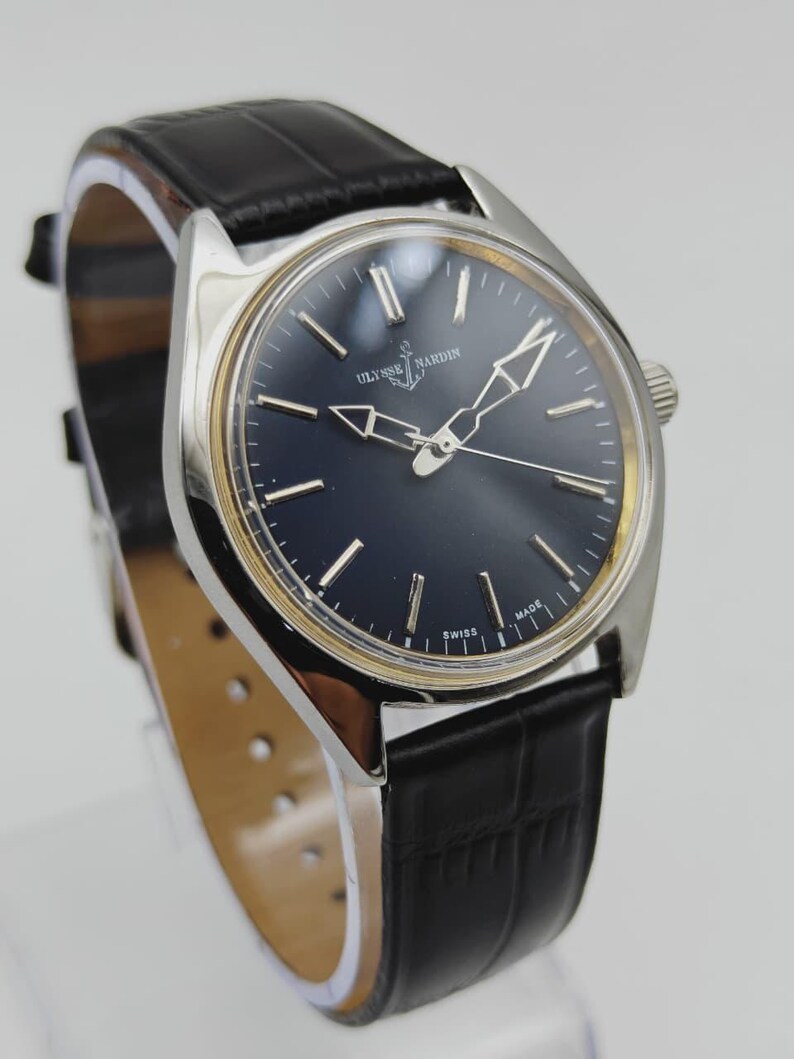 Puede incluir: Un reloj de pulsera Ulysse Nardin plateado con esfera azul oscuro y correa de cuero negro. El reloj presenta agujas e indicadores plateados, con las palabras "SWISS MADE" visibles en la esfera.