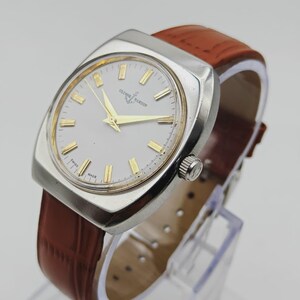 K&ouml;nnte beinhalten: Eine Vintage-Armbanduhr von Ulysse Nardin mit einem silberfarbenen quadratischen Geh&auml;use und einem wei&szlig;en Zifferblatt. Die Uhr hat goldfarbene Zeiger und Markierungen sowie ein braunes Lederarmband. Die Uhr wird auf einem transparenten St&auml;nder ausgestellt.