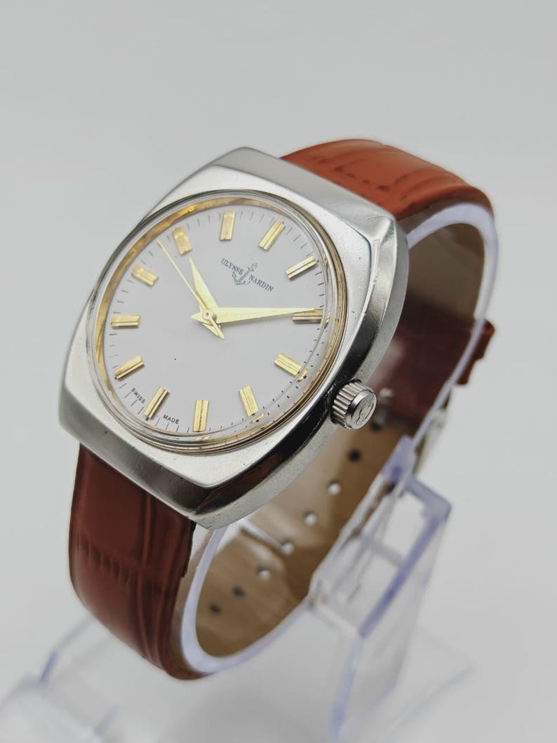 K&ouml;nnte beinhalten: Eine Vintage-Armbanduhr von Ulysse Nardin mit einem silberfarbenen quadratischen Geh&auml;use und einem wei&szlig;en Zifferblatt. Die Uhr hat goldfarbene Zeiger und Markierungen sowie ein braunes Lederarmband. Die Uhr wird auf einem transparenten St&auml;nder ausgestellt.